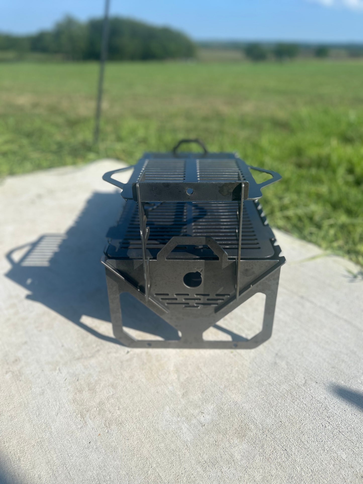 Portable metal grill on a concrete surface with a grassy field in the background